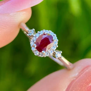 May include: A silver ring featuring a central oval-cut ruby surrounded by a halo of small, clear gemstones. Additional small gemstones are set on the band. The ring is held against a blurred green background.