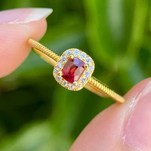 May include: A gold ring featuring a central rectangular red gemstone surrounded by a halo of small white stones. The ring band has a textured, rope-like design along the sides, leading up to the prong-set center stone and its surrounding halo.