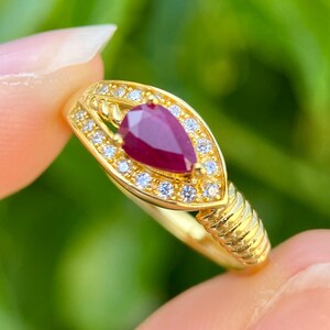 May include: A gold ring with a pear-shaped, deep red gemstone, encircled by small, clear accent stones. The ring's band has a textured design. The ring is held against a blurred green background.