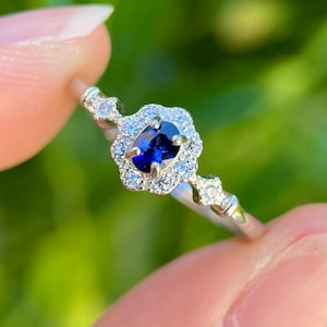 May include: A silver-toned ring featuring a central oval-cut deep blue sapphire stone. The sapphire is surrounded by a scalloped halo of small clear stones. The band includes two additional small clear stones on each side of the central setting.