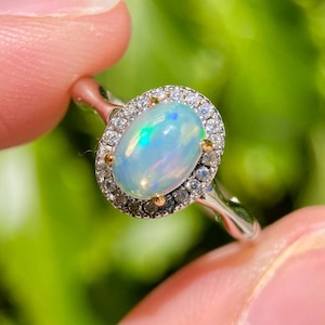 May include: A silver ring featuring a central oval-shaped iridescent opal gemstone. The opal is surrounded by a halo of small, clear, round-cut stones set in a metallic frame. The band is smooth and polished, showcasing a bright, reflective finish.