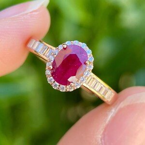 Puede incluir: Un anillo de oro con un rubí rojo intenso de talla ovalada en el centro, rodeado por un halo de pequeños diamantes blancos redondos, con diamantes blancos adicionales de talla baguette engastados a los lados del aro de oro.