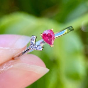 May include: A silver-toned open ring featuring a pear-shaped red gemstone on one end and a letter L shaped accent encrusted with small clear stones on the other end. The band is thin and polished, creating a delicate and refined appearance.