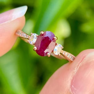 Puede incluir: Anillo de oro rosa con un rubí central de corte ovalado flanqueado por dos piedras claras redondas más pequeñas. La banda está adornada con pequeñas piedras claras. El anillo se sostiene sobre un fondo verde borroso.