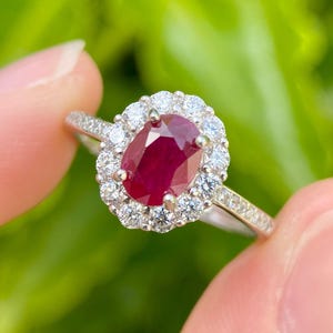 May include: A silver ring with a central oval ruby, encircled by a halo of smaller white diamonds. The ring's band is also set with small diamonds. The background is a blurred green.