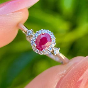May include: A silver ring featuring a large oval-cut ruby gemstone, surrounded by a halo of smaller white gemstones. Two smaller white gemstones flank the ruby. The ring is held up against a blurred green background.
