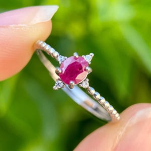 May include: A silver ring featuring a vibrant oval-cut ruby gemstone, accented with small, sparkling white gemstones. The ring's band has a delicate beaded design. The ring is held up against a blurred green background.