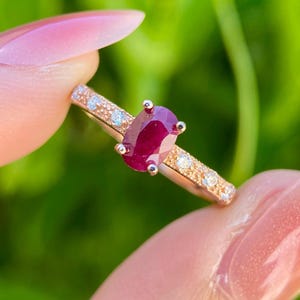 Könnte beinhalten: Ein roségoldener Ring mit einem großen, oval geschliffenen Rubin. Das Band ist mit kleinen, runden, klaren Edelsteinen besetzt. Der Ring wird vor einem verschwommenen grünen Hintergrund gehalten, um das Funkeln und die Farbe des Schmucks hervorzuheben.