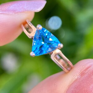 May include: A gold-toned ring featuring a vibrant, triangular, sky-blue gemstone. The ring's band has a simple, elegant design, and the gemstone is the focal point. The background is a soft, blurred green, suggesting an outdoor setting.