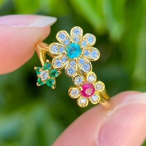 Op de afbeelding: Een gouden ring met drie bloemvormige motieven. De grootste bloem heeft een centrale blauwe steen omringd door heldere kristallen. Een kleinere bloem heeft een rode centrale steen en de derde een groene centrale steen, beide met heldere kristallen.