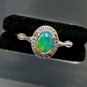 May include: A silver ring featuring an oval-shaped cabochon opal with vibrant green and blue flashes, set in a gold-toned prong setting, surrounded by a halo of small clear round-cut stones on a polished silver band.