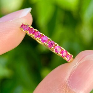 Puede incluir: Un anillo de oro con una fila de pequeñas piedras preciosas de color rosa brillante. El anillo se sostiene entre dos dedos sobre un fondo verde borroso. Las piedras preciosas están espaciadas uniformemente y reflejan la luz.