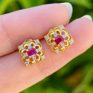 Puede incluir: Un par de pendientes cuadrados dorados. Cada pendiente presenta una gema roja central rodeada de pequeñas gemas transparentes. Los pendientes tienen un diseño calado con un patrón floral. El fondo es verde borroso.