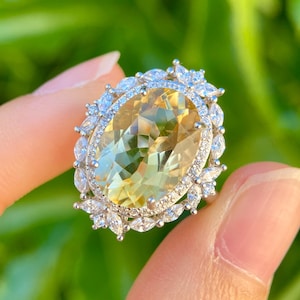 May include: A large, oval-cut yellow gemstone ring with a halo of small, clear stones and decorative accents. The ring is held up against a blurred green background, showcasing the ring's intricate design and color.