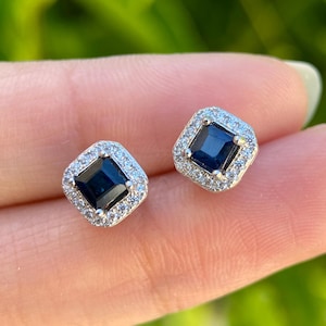 May include: A pair of square-cut, dark blue gemstone stud earrings. Each earring is surrounded by a halo of smaller, clear gemstones, set in a silver-colored metal. The earrings are displayed against a blurred green background.
