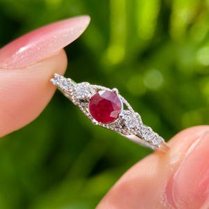 May include: A delicate silver ring featuring a vibrant, round ruby gemstone as the centerpiece. The ring is accented with smaller, sparkling clear stones, creating a refined and elegant design. The ring is held up against a blurred green background.