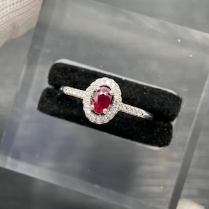 May include: A silver-toned ring featuring a central oval-cut red gemstone surrounded by a halo of small clear stones. The band is slender and also accented with small clear stones along the sides, presented in a black velvet display slot.