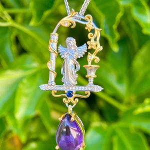 May include: A silver and gold pendant necklace featuring an angel figure within an ornate frame. The pendant includes a large, teardrop-shaped purple gemstone. The necklace hangs on a silver chain, set against a blurred green background.