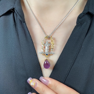 May include: A silver necklace with a pendant featuring a figure within a decorative frame, accented with gold and blue gemstones, and a large, teardrop-shaped purple gemstone. The necklace is worn against a black shirt.