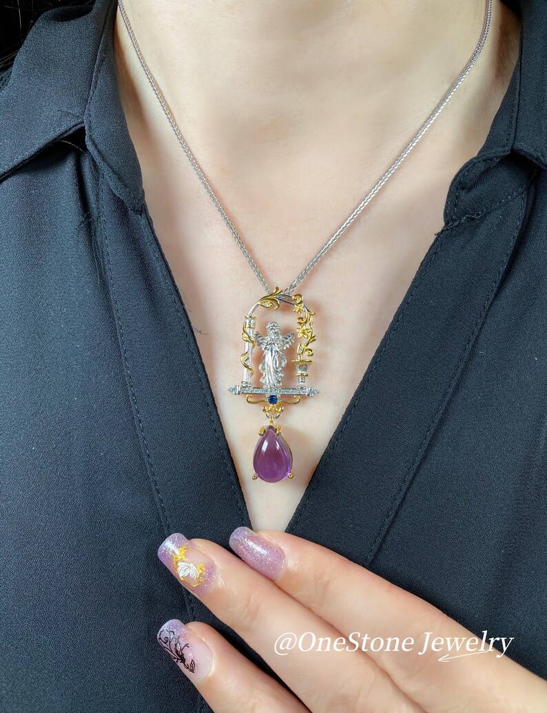May include: A silver necklace with a pendant featuring a figure within a decorative frame, accented with gold and blue gemstones, and a large, teardrop-shaped purple gemstone. The necklace is worn against a black shirt.