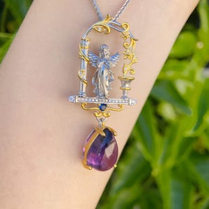 May include: Silver and gold pendant necklace with an angel figure in an ornate frame, and a teardrop-shaped purple gemstone. The necklace hangs from a delicate chain. The text "@OneStone Jewelry" is visible.