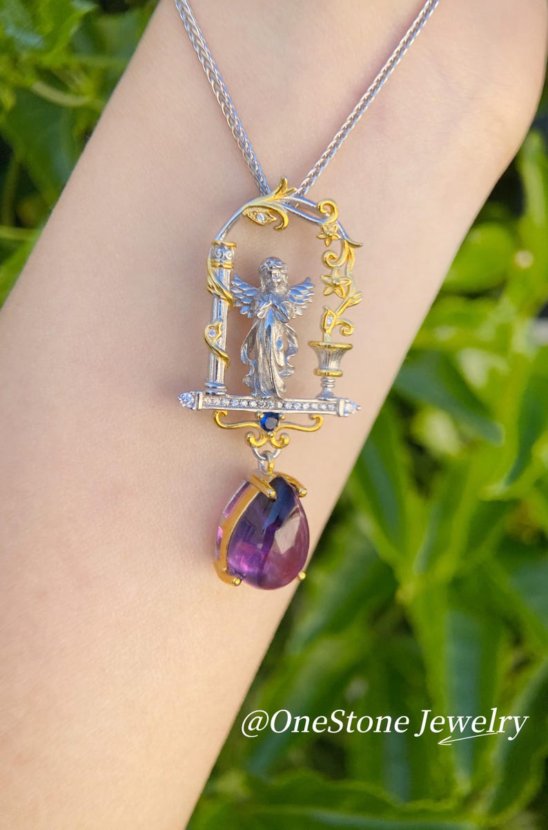 May include: Silver and gold pendant necklace with an angel figure in an ornate frame, and a teardrop-shaped purple gemstone. The necklace hangs from a delicate chain. The text "@OneStone Jewelry" is visible.