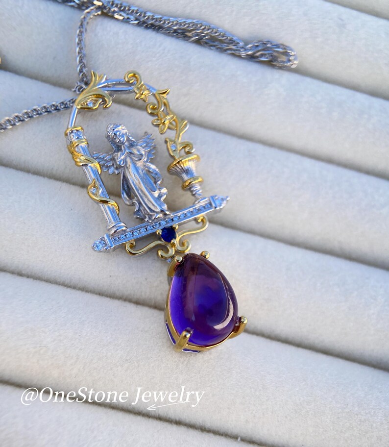 May include: A silver and gold pendant necklace with an angel figure in a decorative frame. A large, teardrop-shaped, deep purple gemstone hangs below. The necklace has a fine silver chain. The text "@OneStone Jewelry" is visible.
