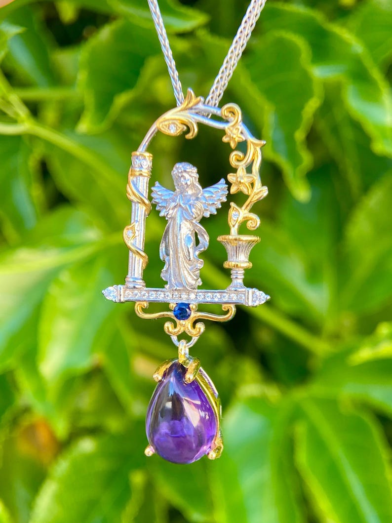 May include: A silver and gold pendant necklace featuring an angel figure within an ornate frame. The pendant includes a large, teardrop-shaped purple gemstone. The necklace hangs on a silver chain, set against a blurred green background.