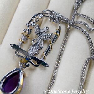 May include: A silver pendant necklace with an angel motif, gold detailing, and a teardrop-shaped purple gemstone. The pendant hangs from a fine silver chain. The necklace showcases an intricate, decorative design.