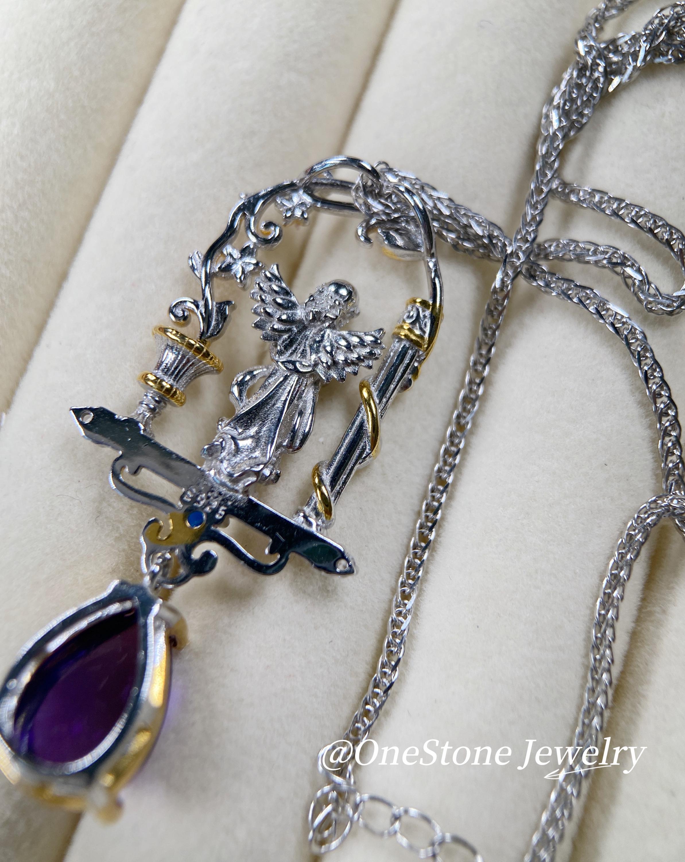 May include: A silver pendant necklace with an angel motif, gold detailing, and a teardrop-shaped purple gemstone. The pendant hangs from a fine silver chain. The necklace showcases an intricate, decorative design.