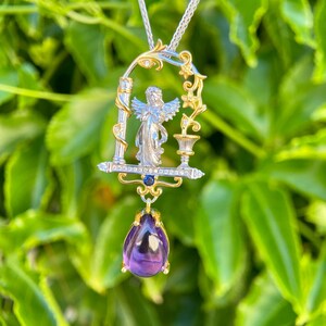 May include: A silver and gold pendant necklace with an angel figure in an arched frame. The pendant has a teardrop-shaped, deep purple amethyst gemstone. The necklace is set against a backdrop of green foliage.
