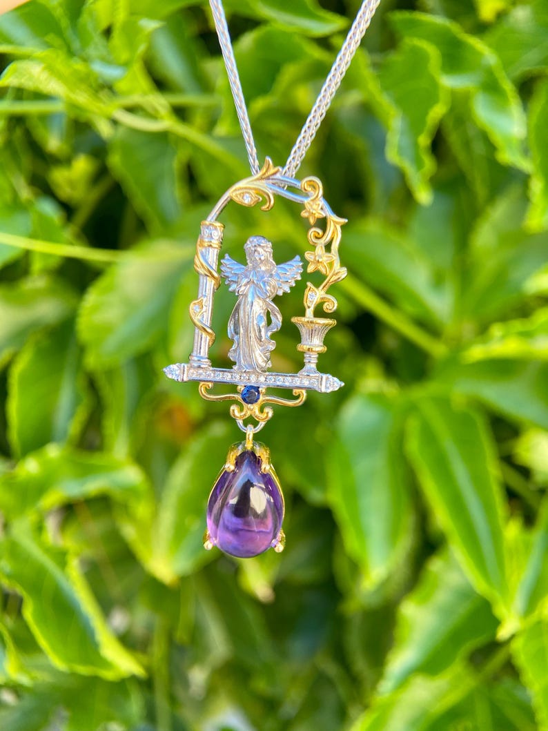 May include: A silver and gold pendant necklace with an angel figure in an arched frame. The pendant has a teardrop-shaped, deep purple amethyst gemstone. The necklace is set against a backdrop of green foliage.