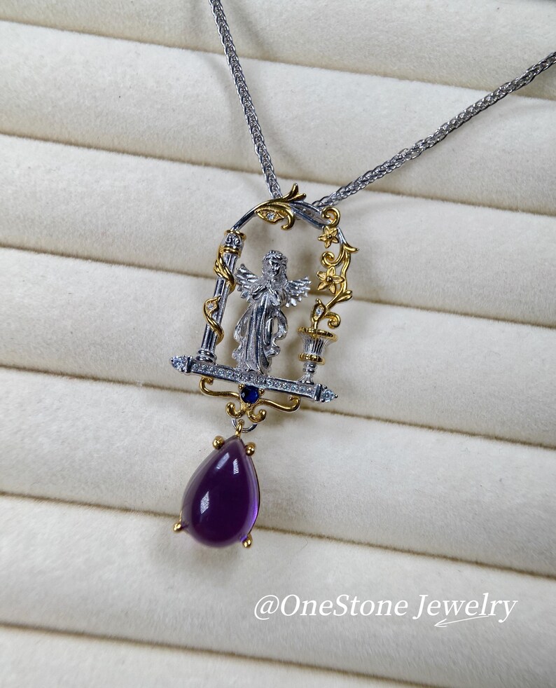 May include: A silver and gold pendant necklace with an angel figure in an ornate frame. The pendant has a large, teardrop-shaped purple gemstone. The necklace is displayed on a white, textured surface. The text "@OneStone Jewelry" is visible.