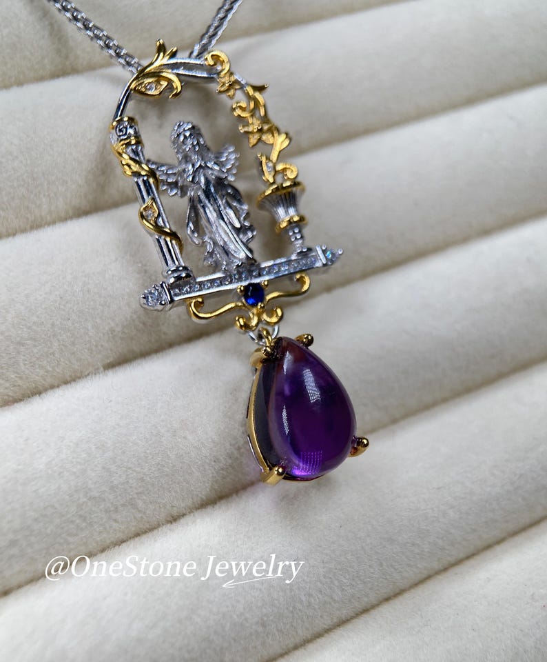 May include: A silver and gold pendant necklace with an angel figure in a decorative frame. The pendant has a large, teardrop-shaped, deep purple amethyst gemstone. The necklace is marked with the text "@OneStone Jewelry".
