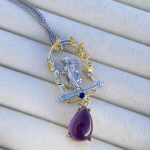 May include: A silver and gold pendant necklace with an angel figure in a decorative frame, blue gemstones, and a teardrop-shaped purple amethyst. The necklace hangs from a fine chain. The text "@OneStone Jewelry" is visible. The measurements are in cm.