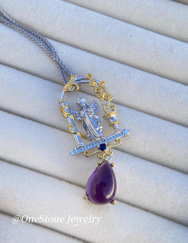 May include: A silver and gold pendant necklace with an angel figure in a decorative frame, blue gemstones, and a teardrop-shaped purple amethyst. The necklace hangs from a fine chain. The text "@OneStone Jewelry" is visible. The measurements are in cm.