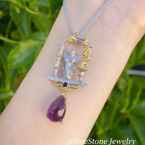 May include: A silver and gold pendant necklace with an angel design, set within an ornate frame, and a teardrop-shaped purple gemstone. The necklace has a fine chain and is set against a blurred green backdrop. The text "@OneStone Jewelry" is visible.