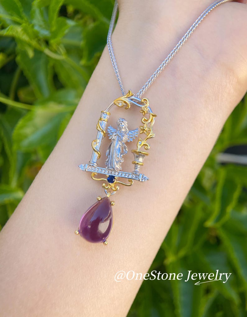 May include: A silver and gold pendant necklace with an angel design, set within an ornate frame, and a teardrop-shaped purple gemstone. The necklace has a fine chain and is set against a blurred green backdrop. The text "@OneStone Jewelry" is visible.