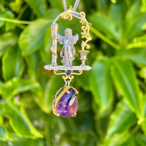 May include: A silver pendant necklace with an angel figure in a gold-accented arched frame. The pendant features a teardrop-shaped, purple amethyst gemstone. The necklace is set against a backdrop of green leaves.