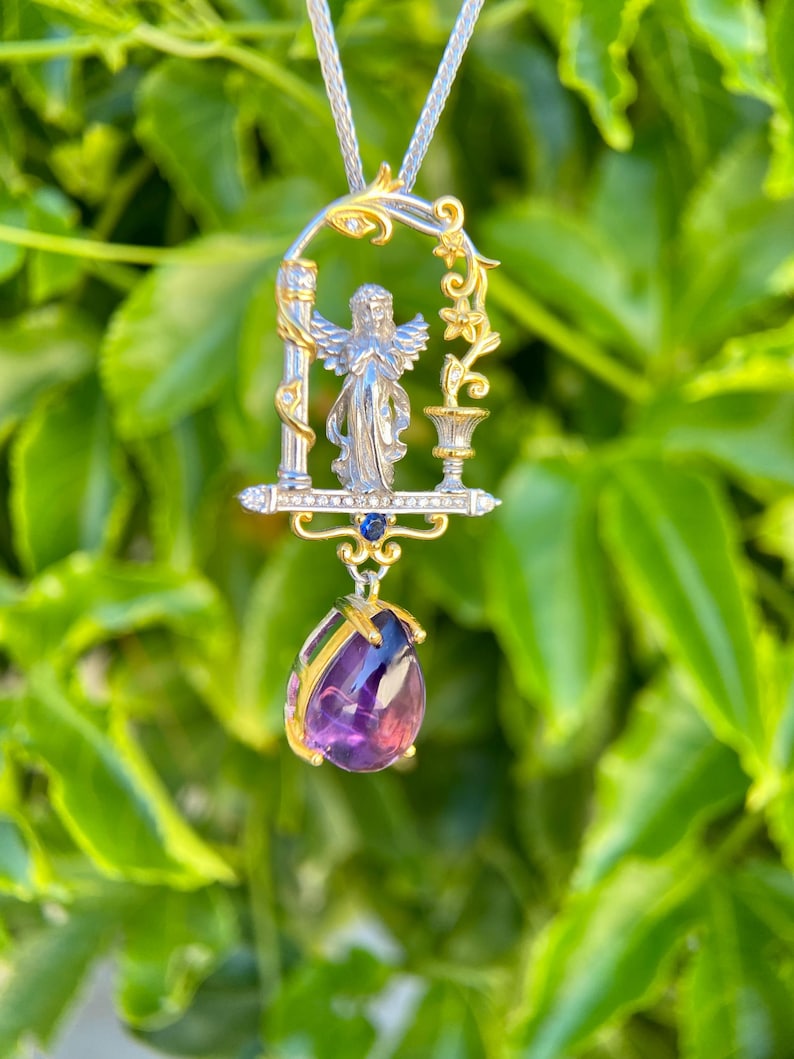 May include: A silver pendant necklace with an angel figure in a gold-accented arched frame. The pendant features a teardrop-shaped, purple amethyst gemstone. The necklace is set against a backdrop of green leaves.