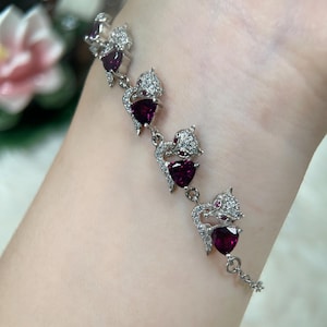 May include: A silver bracelet featuring a series of stylized cat figures. Each cat holds a heart-shaped, deep red gemstone. The cats are embellished with small, clear crystals. The bracelet is delicate and elegant, with a chain closure.