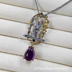 May include: A silver and gold pendant necklace with an angel figure in an ornate frame, set with small diamonds and a deep purple, teardrop-shaped gemstone. The necklace has a fine chain and is set against a soft, textured background.