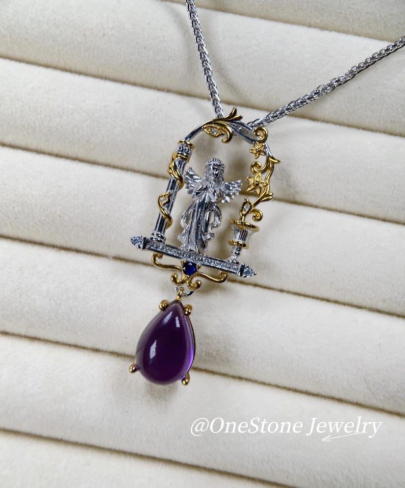 May include: A silver and gold pendant necklace with an angel figure in an ornate frame, set with small diamonds and a deep purple, teardrop-shaped gemstone. The necklace has a fine chain and is set against a soft, textured background.