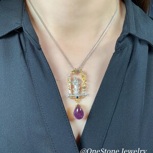 May include: A silver necklace with a gold pendant featuring a figure, clear stones, and a teardrop-shaped purple gemstone. The pendant hangs from a delicate silver chain. The text "@OneStone Jewelry" is at the bottom.