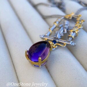 May include: A silver and gold pendant necklace with a large, teardrop-shaped purple gemstone. The pendant has a detailed design with a figure and is suspended from a delicate chain. The text "@OneStone Jewelry" is visible.