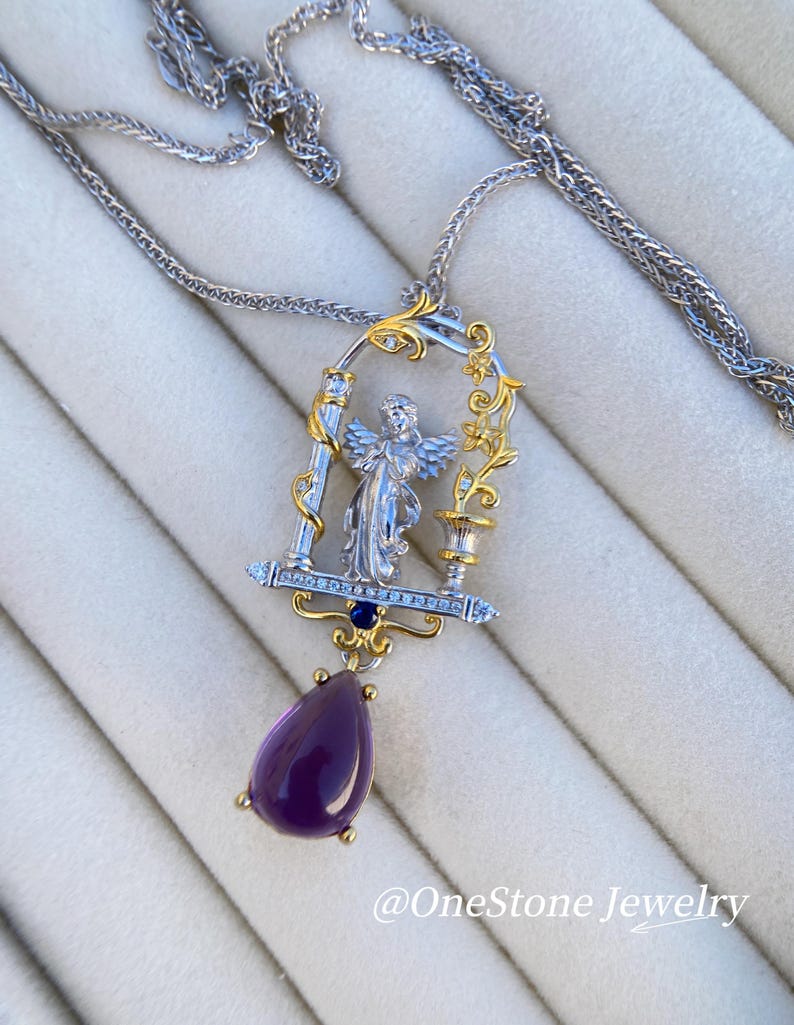 May include: A silver and gold pendant necklace with an angel design. The pendant has a teardrop-shaped purple gemstone and is suspended from a fine chain. The necklace features the text "@OneStone Jewelry".