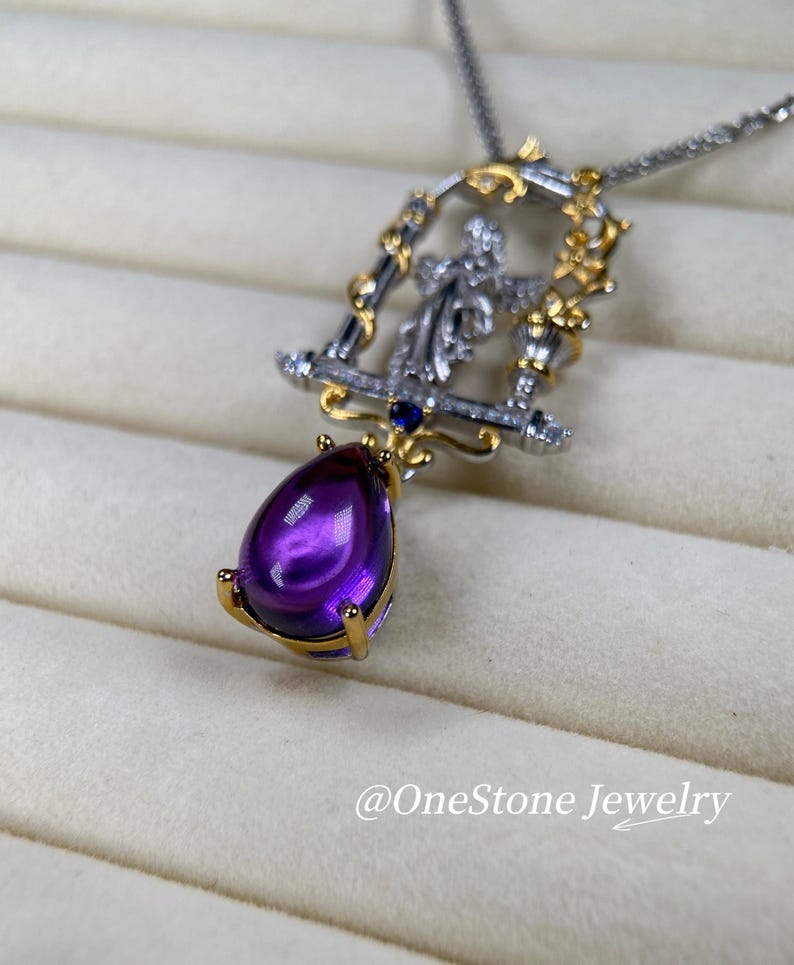 May include: A silver necklace with a pendant featuring a large, teardrop-shaped purple amethyst gemstone. The pendant has a gold-coloured frame with intricate detailing and a small figure. The necklace is displayed on a light-coloured surface. The text "@OneStone Jewelry" is visible.