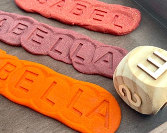 May include: Three strips of glittery modeling dough in red, maroon, and orange, each embossed with the names ISABEL and BELLA. A light-colored wooden cube with the letter E and the number 2 visible on its sides sits next to the dough strips.