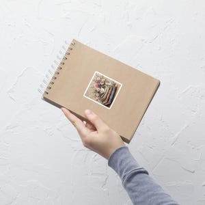 Könnte beinhalten: Ein Aufkleber mit einer Aquarellillustration eines Blumenstraußes und Büchern. Die Blumen sind rosa, weiß und lila, und die Bücher haben verschiedenfarbige Buchrücken. Der Aufkleber befindet sich auf einem braunen Spiralnotizbuch.