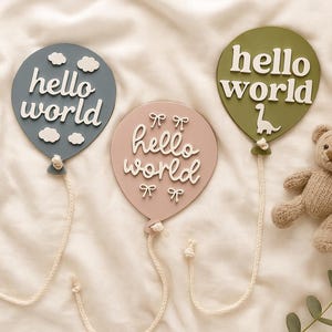 以下が含まれることがあります： 青、ピンク、緑の3つの木製風船型飾りで、それぞれに白い文字で「hello world」と書かれています。小さなテディベアと、「B」と「E」の文字が入った木製ブロックもあります。赤ちゃん用の飾りです。